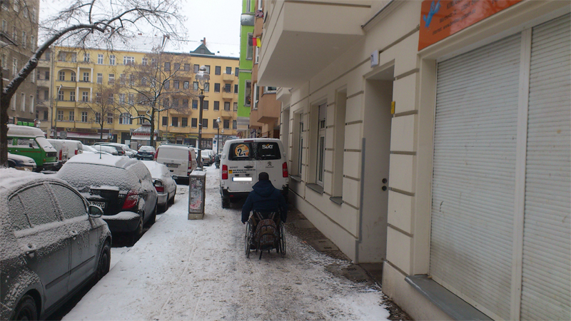 Rollstuhlfahrer vor einem auf dem Gehweg geparkten Mietauto, das ihm die Weiterfahrt versperrt