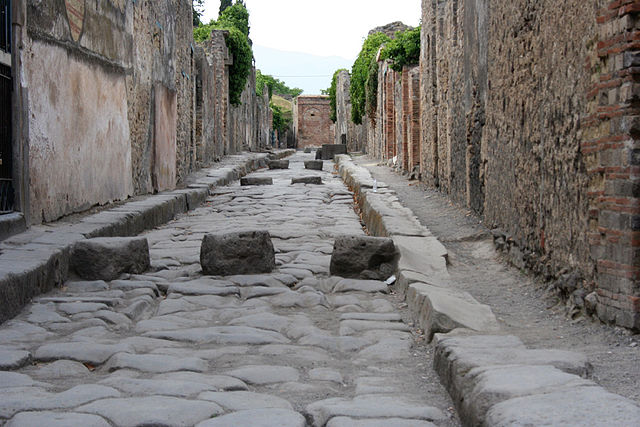 Große Steinquader liegen in einer Reihe quer zur Straße, im Hintergrund eine antike Stadtreste